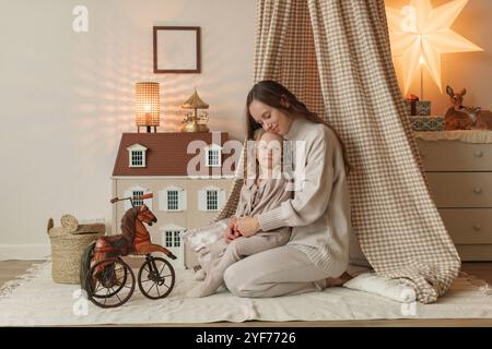 Happy mother and daughter sitting in a play teepee next to a doll's house in a bedroom Stock Photo