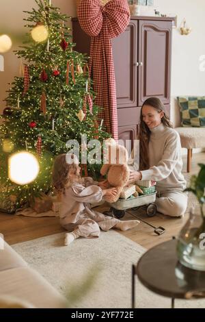 Smiling Mother and daughter in leisurewear sitting on the floor in a living room next to an illuminated Christmas tree playing with a teddy bear Stock Photo