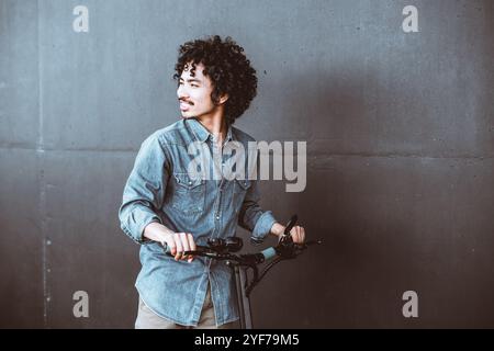 A young asian man with curly hair wearing a casual denim shirt poses confidently while holding an electric scooter against a minimalist dark concrete Stock Photo