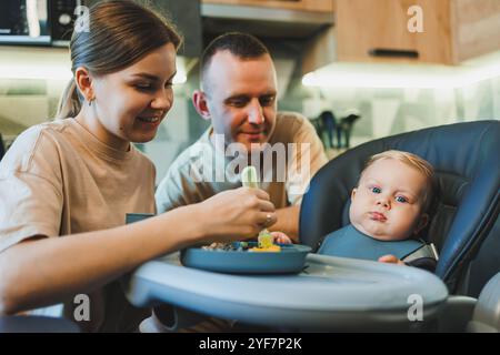 The first complementary food for the baby with a spoon. A mother feeds puree to her 6-month-old son. Parents feed their baby with food on the bed Stock Photo