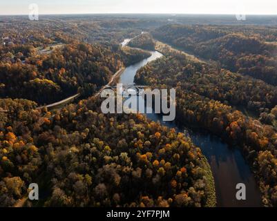 An aerial view captures a winding river and bridge amidst vibrant autumn foliage in Sigulda, Latvia, with hills and valleys in the background. Stock Photo