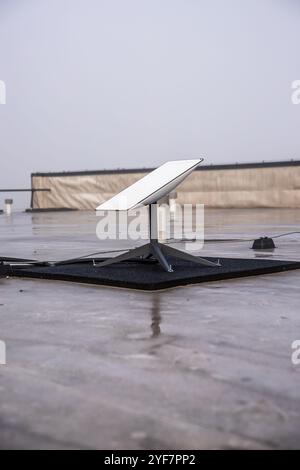 A Starlink satellite dish is mounted on a wet rooftop, with cables extending from it. The overcast sky and low wall suggest an urban environment. Stock Photo