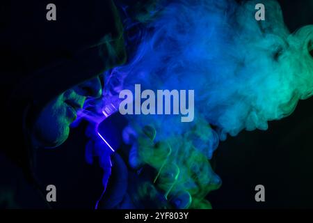 male vaper holding vape electronic cigarette close-up on background of colored smoke, concept of harm of smoking Stock Photo