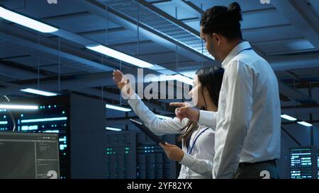 Admins in data center managing and maintaining databases using device, ensuring data integrity and security. IT professionals updating server room workspace systems using tablet Stock Photo