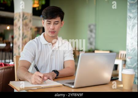 A serious, focused Asian man is working remotely from a coffee shop, sitting at an indoor table with his laptop and taking notes in his notebook. peop Stock Photo