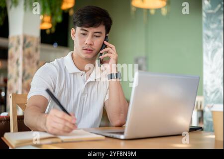 A focused Asian man is working remotely from a coffee shop, sitting at an indoor table with his laptop and talking on the phone while taking notes in Stock Photo