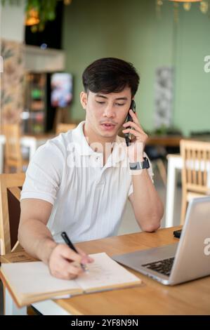 A focused Asian man is working remotely from a coffee shop, sitting at an indoor table with his laptop and talking on the phone while taking notes in Stock Photo