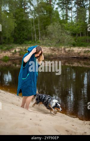Young Woman in Blue Poncho Relaxing by Forest River, Curious Dog Exploring the Calm Waters Stock Photo