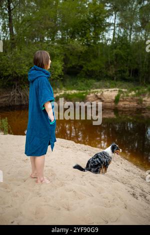 Young Woman in Blue Poncho Relaxing by Forest River, Curious Dog Exploring the Calm Waters Stock Photo
