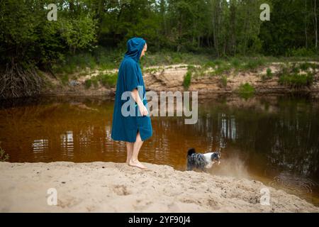 Man in Teal Poncho Standing by the River, Dog Playing by the Riverside Stock Photo