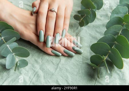 Beautifully manicured hands showcase long, green nails adorned with a delicate ring. The setting features soft eucalyptus leaves, creating a serene am Stock Photo