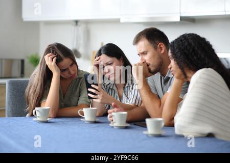 Four sad interracial friends checking mobile phone and complaining at home Stock Photo
