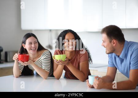 Three interracial friends talking and drinking coffee or tea  in the kitchen at home Stock Photo