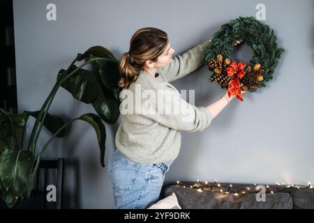 Woman hanging handmade Christmas wreath with pinecones and red ribbon. DIY holiday decor adds a personal touch to festive home decorating. Stock Photo