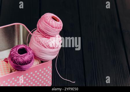 Assorted yarn and sewing tools in a pink tin box on a black wooden background create a visually appealing arrangement for craft lovers. Stock Photo