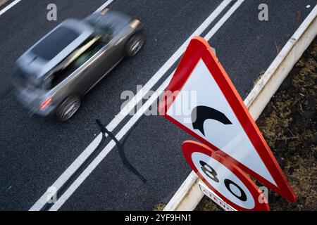 A gray car moves along the road next to a huge caution and speed limit sign. The ground is wet from the rain and there is a lot of contrast between bl Stock Photo