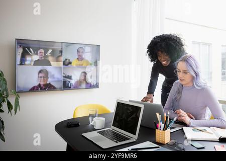 Smiling businesswoman discussing with female colleague while attending video conference through television set in office Stock Photo