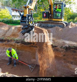 Man in tractor pouring dirt in trench while coworker using pitchfork standing in trench Stock Photo