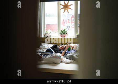 Smiling boy lying beside non-binary sibling using smart phone while lying on bed at home Stock Photo