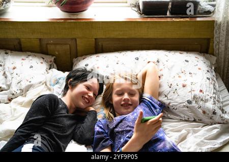 High angle view of happy boy lying beside non-binary sibling using smart phone while lying on bed at home Stock Photo