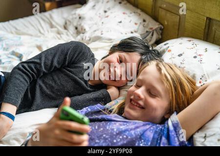 Excited boy lying beside non-binary sibling using smart phone while lying on bed at home Stock Photo