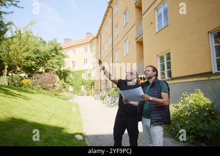 Two male architects examining residential building while holding blueprint and standing on footpath at sunny day Stock Photo