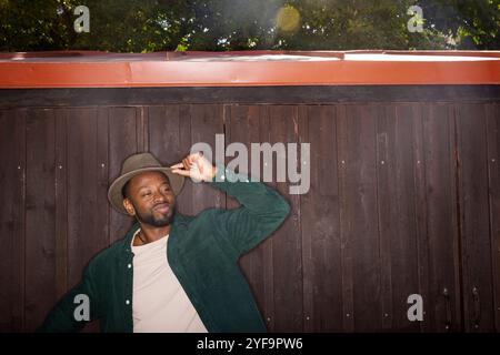 Smiling young man tipping hat while leaning on wall Stock Photo
