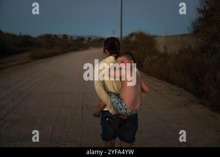 A 3-year-old boy rubs his eyes carried on his mom's back on dirt road Stock Photo