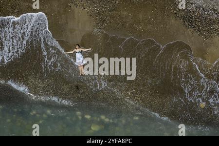 Woman with swimsuit and white dress resting on a sandy beach with breaking waves on the shore. Overhead shot. Aerial drone photograph Stock Photo