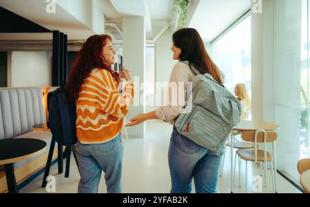 Two friends are seen chatting together on campus, walking in a modern university cafe with backpacks. The scene captures a moment of youthful energy a Stock Photo