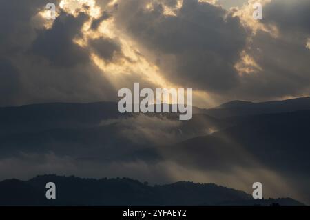 Bright sun rays light shining through dark clouds over mountain at sunset. Dramatic sky in winter Stock Photo