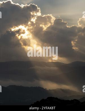 Bright sun rays light shining through dark clouds over mountain at sunset. Dramatic sky in winter Stock Photo