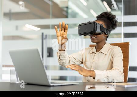 Young african american business woman using VR headset in modern office. Embracing technology, interacting with virtual reality, and exploring innovative solutions. Concept future work environments. Stock Photo