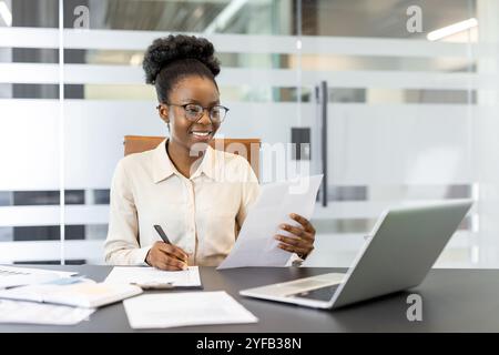 Young African American businesswoman examining papers at office desk with laptop, pens, and documents. Portrays professional work environment, focus, and productivity. Stock Photo