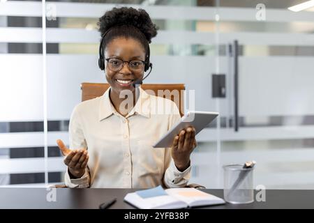Young African American businesswoman with headset using tablet for virtual meeting in modern office. Smiling, confident expression. Ideal for concepts of communication, customer support, remote work. Stock Photo