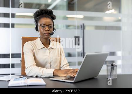 Young African American business woman in modern office wearing headset, using laptop for professional call. Focused on digital communication and customer service tasks. Stock Photo