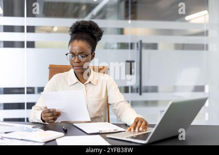 Young african american business woman focused on reading documents at desk in modern office. Professional environment with laptop and paperwork showcasing concentration and productivity. Stock Photo