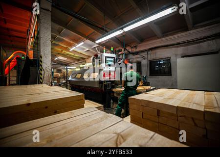 A worker operates a computerized machine in a woodworking factory, processing large wooden beams in an industrial setting Stock Photo