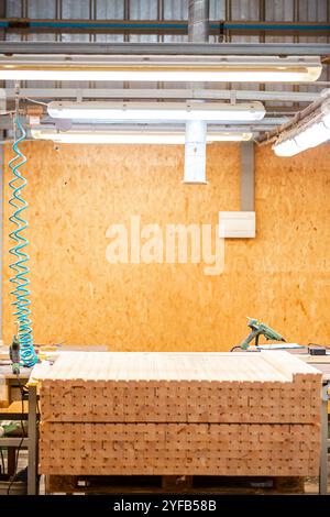 workshop table with stacked wooden planks, tools, and equipment, under bright fluorescent lights against a wooden wall backdrop in an industrial setti Stock Photo