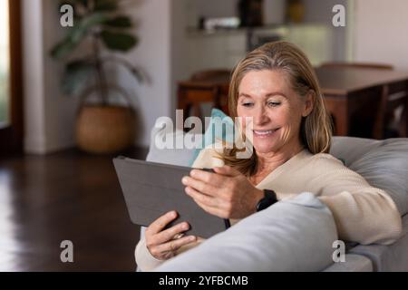 Smiling senior woman using tablet at home, relaxing on cozy sofa Stock Photo