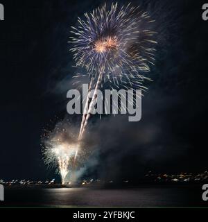 Fireworks exploding over water at night with reflections and smoke during a public celebration event. Stock Photo