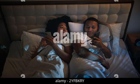 African American couple in bed at night late time in game bedroom relaxing using two phones chatting in mobile app scrolling social media smartphones Stock Photo