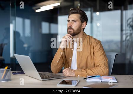Mature businessman in modern office environment sitting at desk with laptop, phone, and open notebook, conveying thoughtful expression. Professional setting highlights business and work. Stock Photo