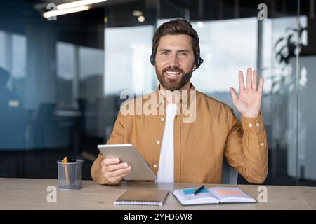 Mature businessman with headset using tablet, waving during virtual meeting in office. Confident and friendly expression, showcasing business communication and technology use. Stock Photo