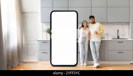 A couple stands happily in a modern kitchen next to a large blank phone screen Stock Photo