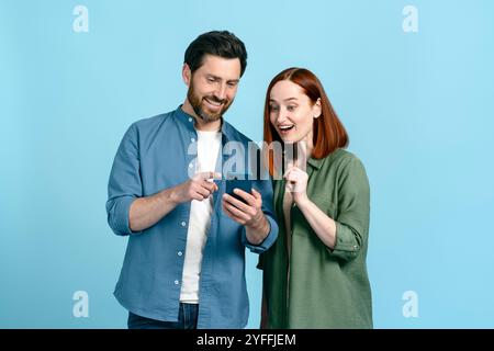 Young man and a woman are looking at smartphone screen with excitement, posing on blue studio background Stock Photo
