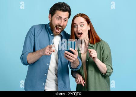 Young man and woman are holding a smartphone and pointing at it with surprised expressions on their faces Stock Photo