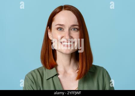 Joyful young woman with red hair smiles brightly against a blue background, exuding confidence and approachability Stock Photo