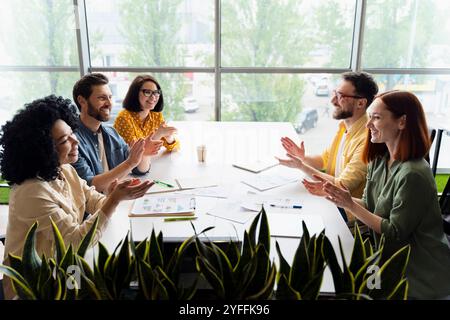 Diverse team of coworkers is sitting around a table with documents and computers, clapping their hands Stock Photo