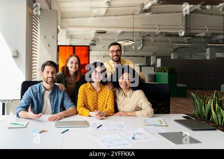 Team of five professionals is smiling while sitting at a table in their office, working together on a project Stock Photo